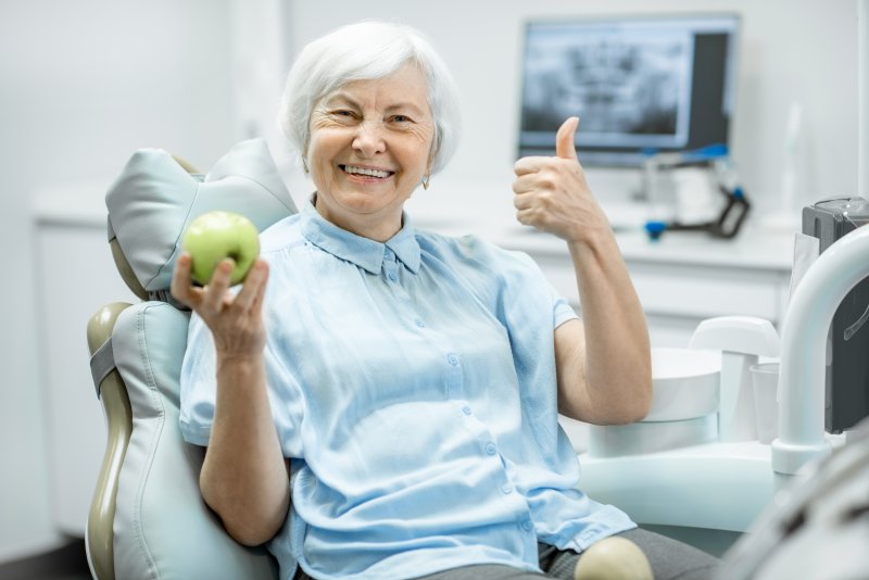 A smiling senior woman giving a thumbs up, holding a green apple, and smiling in a dentist’s chair